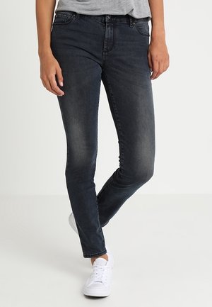 Personne portant un jean slim foncé et des baskets blanches, debout avec une jambe légèrement pliée sur un fond clair uni.