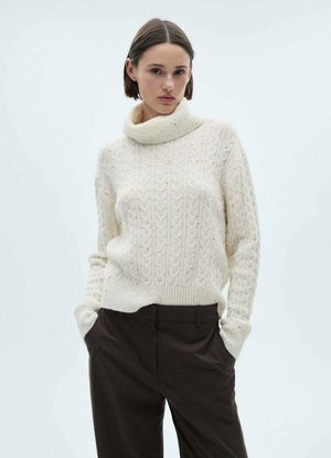 Cremfarbener gestrickter Pullover mit hohem Kragen, der ein strukturiertes Zopfmuster und gerippte Bündchen aufweist, kombiniert mit dunkelbraunen Hosen.