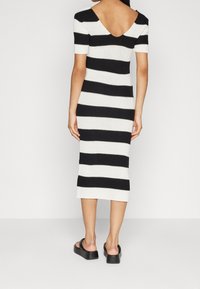 Figurbetontes Strickkleid mit kurzen Ärmeln, das abwechselnd schwarze und weiße Streifen zeigt und einen weiten V-Ausschnitt aufweist, der bis zur Mitte der Wade reicht. Dazu werden schwarze Sandalen getragen.