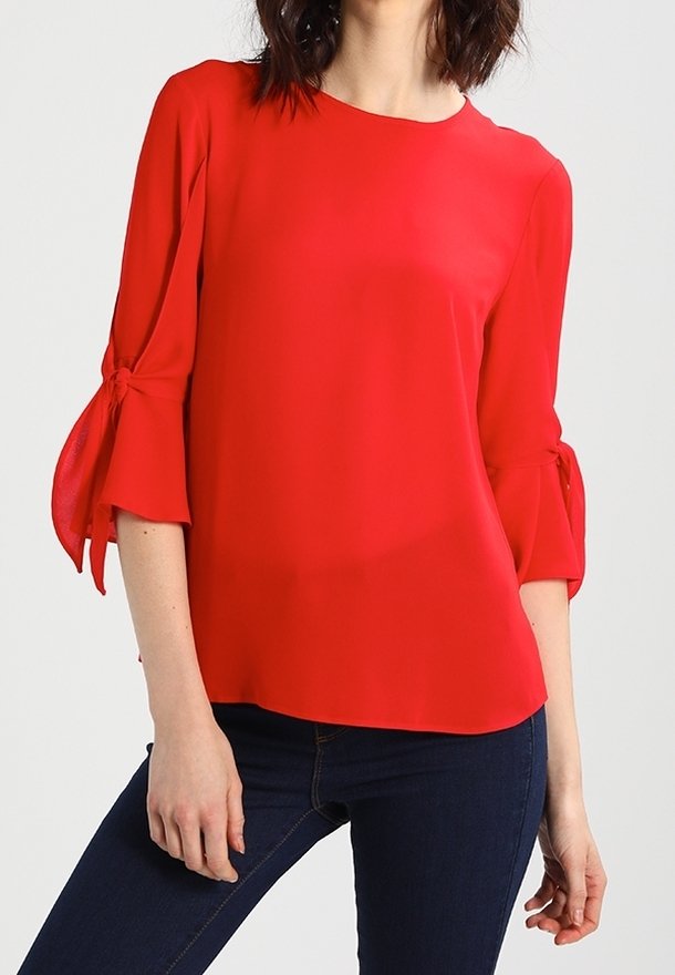 Femme portant un chemisier rouge vif à manches trois-quarts nouées en nœuds, associé à un jean bleu foncé, sur un fond clair uni.