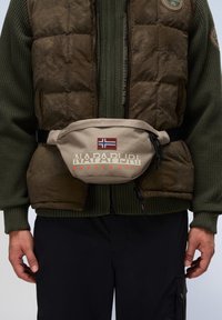 Beige midjeväska med justerbar svart rem, prydd med en norsk flagga-badge och röd "NAPAPIJRI" logotyp med orange text, strukturerad tyg.