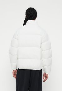 Bijela puffer jakna s visokim ovratnikom, koja ima izraženo horizontalno kviltanje i opušteni kroj. Glatka tekstura, minimalno brendiranje na rubu.
