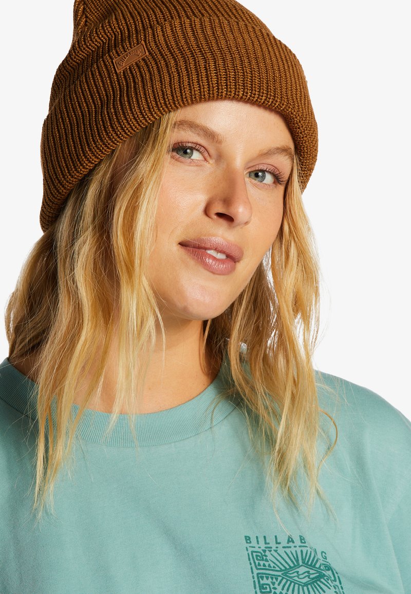 Berretto lavorato a maglia marrone con texture a coste e piccola etichetta logata, abbinato a una maglietta leggera a collo rotondo di colore azzurro con un design grafico.