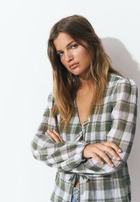 Blouse à carreaux vert et blanc avec un design à nouer à l'avant, manches longues et coupe décontractée ; tissu léger texturé ajoutant de l'intérêt visuel.
