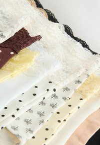 Sous-vêtements pour femmes superposés avec bordure en dentelle, en blanc, noir, marron, jaune et motifs à pois, disposés en diagonale sur une surface claire.