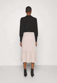 Femme se tenant de dos, portant un haut noir à manches longues, une jupe midi beige avec un ourlet à volants, et des bottines noires à talons.