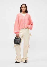 Ροζ oversized πλέκτο με κουμπιά, κρεμ παντελόνι με λεπτομέρειες από κορδόνια και μια μικρή μαύρη τσάντα με ατσάλινες λεπτομέρειες. Flat μαύρα παπούτσια.