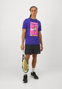 Μωβ μπλούζα Nike με πολύχρωμο γραφικό σχέδιο, συνδυασμένη με μαύρα αθλητικά σορτς και μαύρα αθλητικά παπούτσια, κρατώντας μια ρακέτα τένις.