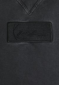 Sudadera negra con una tela texturizada, diseño de cuello en V y un parche rectangular negro bordado con un detalle de firma.