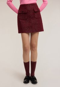 Bordeauxrode minirok met structuur, voorzien van twee klepzakken aan de voorkant en zwarte knopen, gecombineerd met kniekousen met ribbelstructuur en zwarte loafers.