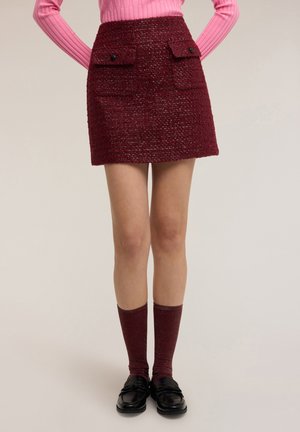 Minifalda burdeos texturizada con dos bolsillos con solapa en el frente y botones negros, combinada con calcetas altas acanaladas hasta la rodilla y mocasines negros.