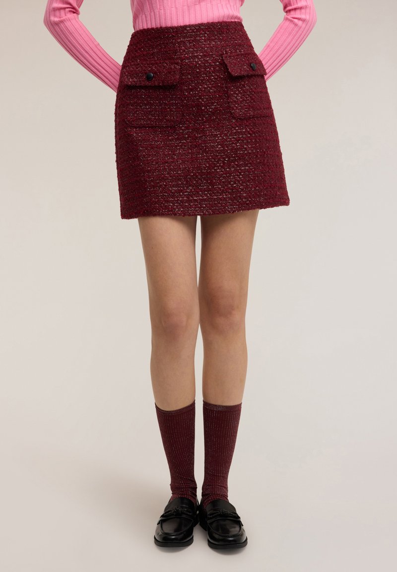 Bordeauxrode minirok met structuur, voorzien van twee klepzakken aan de voorkant en zwarte knopen, gecombineerd met kniekousen met ribbelstructuur en zwarte loafers.