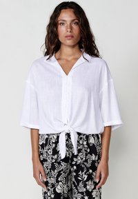 Camicia bianca a maniche corte con bottoni e nodo sul davanti, realizzata in tessuto leggero. Abbinata a pantaloni neri con motivo floreale.