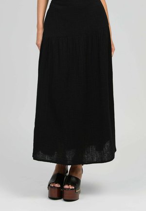 Femme portant une longue jupe plissée noire associée à des talons ouverts à plateformes noirs avec semelles en bois et clous dorés.