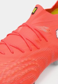 Lyse koralfarvede fodboldsko med tekstureret overdel, tynde snørebånd og forstærket tå. Har et logo på tungen og kontrasterende detaljer.