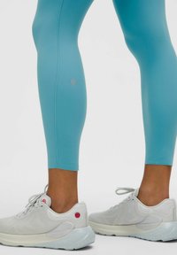 Gai�ši zili augstas veiktspējas legingi ar gludu tekstūru un logotipu pie potītes, apvienoti ar baltām sporta kedām ar nelieliem akcentiem.