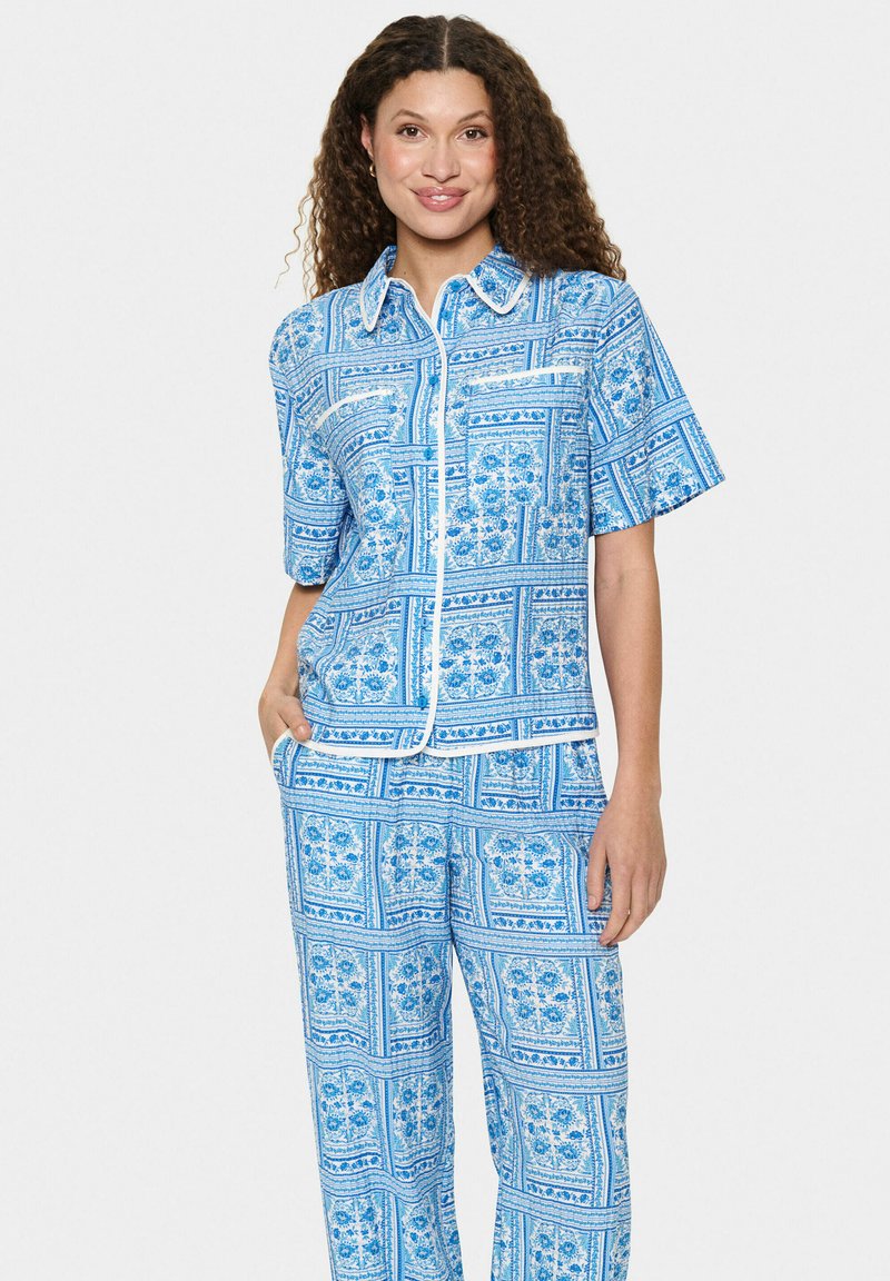 Modrá vzorovaná sada košele a nohavíc s krátkymi rukávmi, zdobená kvetinovými motívmi a kontrastným bielym lemovaním, vyrobená z mäkkej tkaniny.