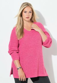 Übergroßer, rosa Strickpullover mit V-Ausschnitt, umgeschlagenen Bündchen, Seitenschlitzen und einer strukturierten Musterung. Kombiniert mit schwarzen Leggings.