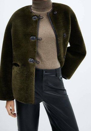 Personne portant une veste duveteuse vert foncé avec des boutons brandebourg, un pull à col roulé beige et un pantalon en cuir noir devant un fond uni.