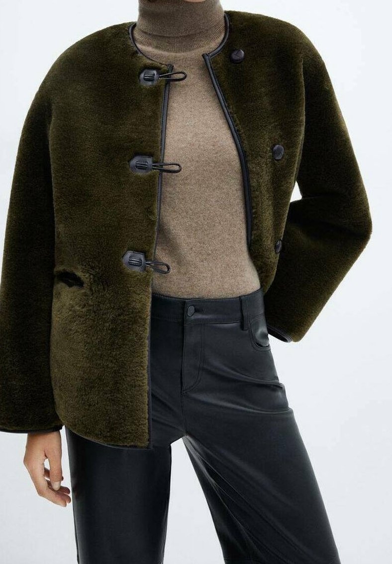 Personne portant une veste duveteuse vert foncé avec des boutons brandebourg, un pull à col roulé beige et un pantalon en cuir noir devant un fond uni.