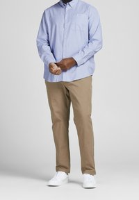 Camicia leggera azzurra con taschino sul petto, abbinata a pantaloni color khaki e sneakers bianche. La camicia ha un colletto classico e maniche lunghe.