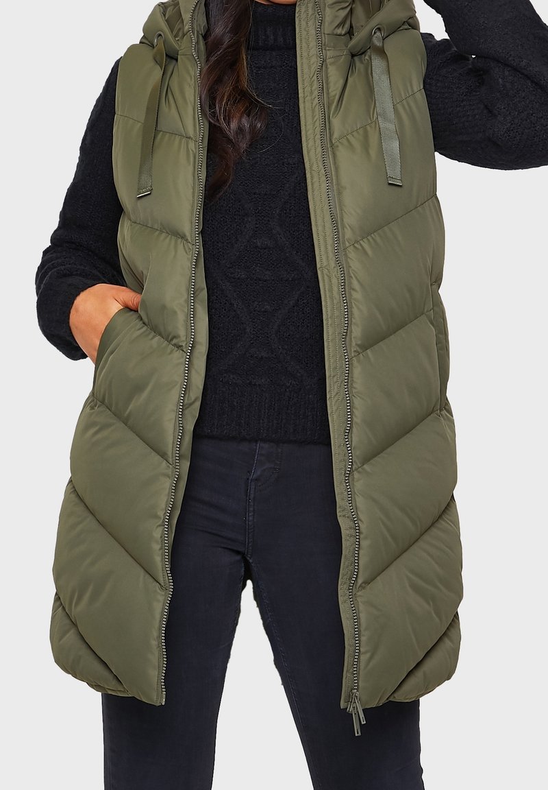 Olive green puffer vest with a hood, featuring diagonal quilting, a front zip closure, and two side pockets. Worn over a black sweater.