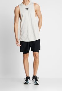 Camiseta sin mangas gris claro con un logo de toro negro, combinada con pantalones cortos deportivos negros y zapatillas negras con acentos en azul verdoso.