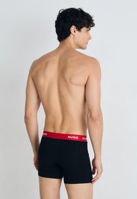Schwarze Boxer-Shorts mit einem roten elastischen Bund, der mit "HUGO" beschriftet ist. Der Stoff scheint glatt und enganliegend zu sein und zeigt ein minimalistisches Design.
