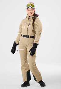 Tuta da sci beige con accenti rosa, cintura e molte cerniere. Guanti e stivali neri. La modella indossa un berretto rosa e occhiali da sci. Sfondo semplice.