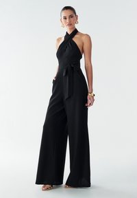 Combinaison noire à encolure halter avec jambes larges, en tissu texturé, détail de taille nouée, comprenant des poches latérales et un design minimaliste.