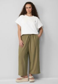 Weißes Kurzarm-T-Shirt mit floralen Stickereien, kombiniert mit olivgrünen weit geschnittenen Hosen und beigen Plattform-Sandalen vor einem neutralen Hintergrund.