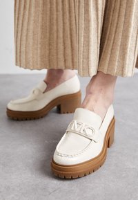Beigefärgade läderloafers med en hög, grov brun plattformssula och ett dekorativt logoband över framsidan. Slät textur och minimalistisk design.