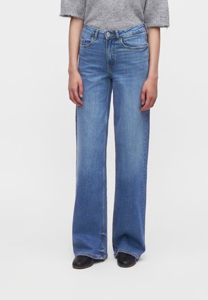 Persoon die lichtblauwe wijde jeans, een grijze korte mouw trui en zwarte puntige schoenen draagt, staand tegen een effen witte achtergrond.