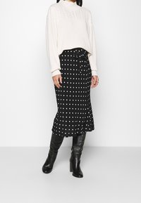 Pull en tricot crème, jupe midi à pois noirs et bottes noires à hauteur de genou. La jupe possède un lien à la taille et un ourlet froncé.