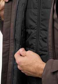 Giacca isolante marrone con tasca interna con zip. Mano che afferra la zip, mostrando un rivestimento nero a contrasto e una texture trapuntata.