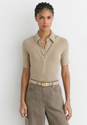 Vrouw met gevlochten haar die een beige korte mouwen hemd met kraag draagt, ingestopt in een hoge bruine broek met een beige riem.