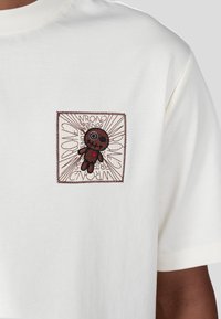 Bílý bavlněný tričko s čtvercovou hnědou vyšívanou nášivkou s karikaturou a textem, umístěné na texturovaném pozadí.