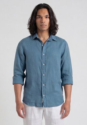 Jeune homme aux cheveux foncés mi-longs, portant une chemise bleue boutonnée aux manches retroussées et un pantalon blanc, debout devant un fond uni.