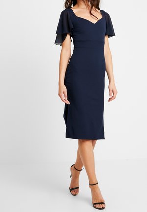 Kobieta ubrana w dopasowaną granatową sukienkę midi z falbanowymi rękawami i czarne czółenka na pasku wokół kostki, stojąca ze skrzyżowanymi nogami.