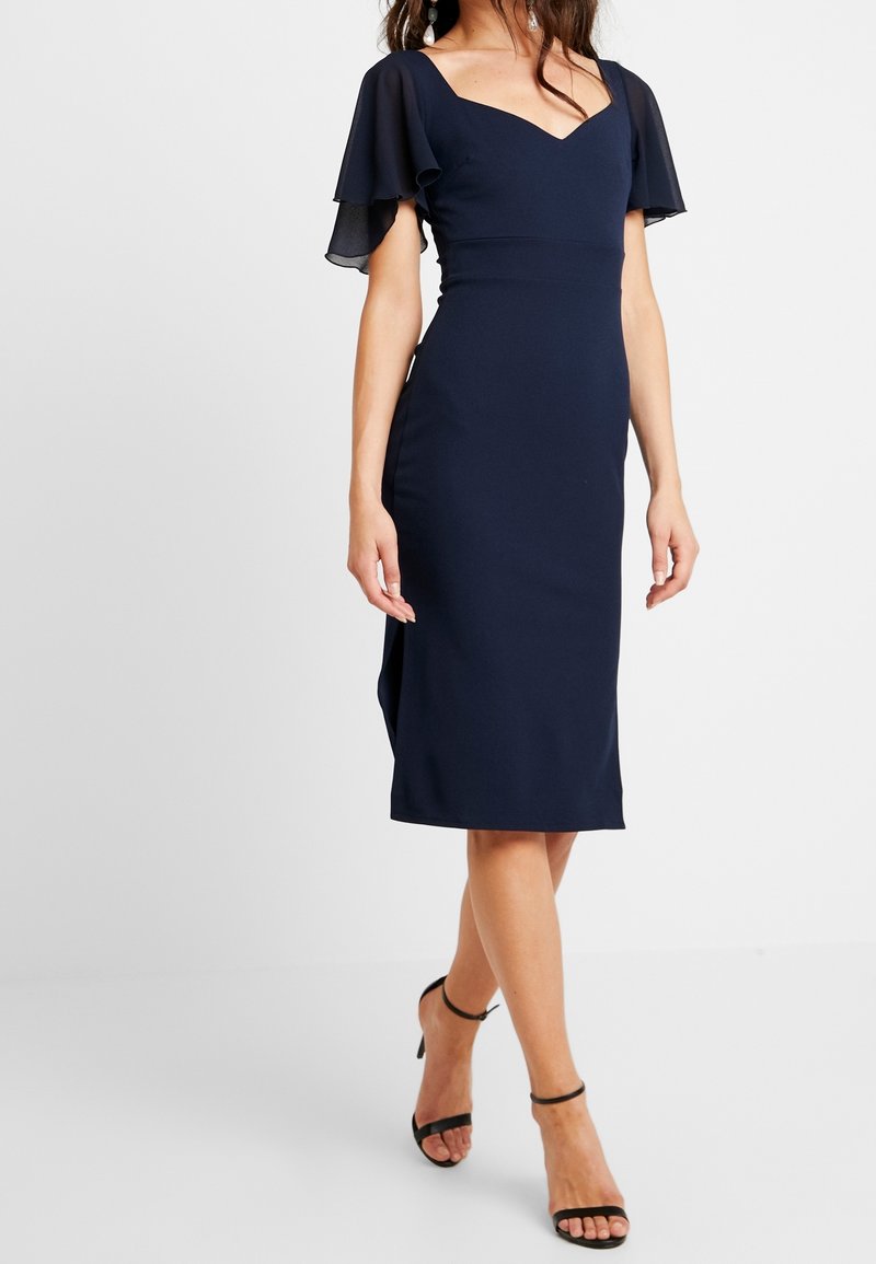 Femme portant une robe midi bleu marine ajustée avec des manches volantées et des talons noirs à bride à la cheville, debout les jambes croisées.
