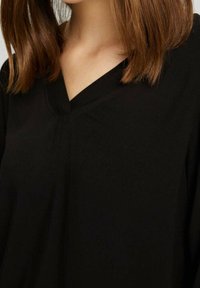 Blusa nera con scollatura a V, caratterizzata da dettagli di cucitura minimi, texture liscia e un tessuto fluido che si drappeggia attorno alle spalle. Design semplice e versatile.