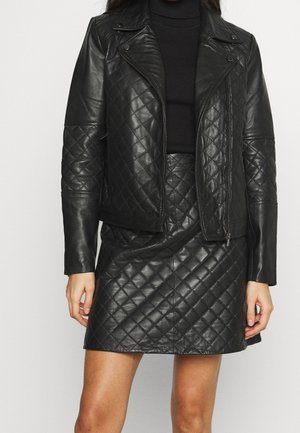 Frau trägt schwarze gesteppte Lederjacke über einem schwarzen Rollkragenpullover und einem passenden gesteppte Lederrock.