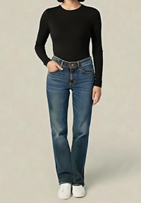 Personne portant un haut à manches longues noir, un jean taille haute bleu, des chaussures blanches sans lacets, avec une main dans la poche et un fond neutre.