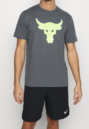 Mężczyzna w ciemnoszarej koszulce z neonowym logo byka na głowie i czarnych spodenkach Nike, stojący na jasnym tle.