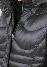 Doudoune gris foncé, à motif matelassé, avec une capuche bordée de fourrure et une fermeture éclair avant ornée de boutons-pression. Texture douce et brillante.