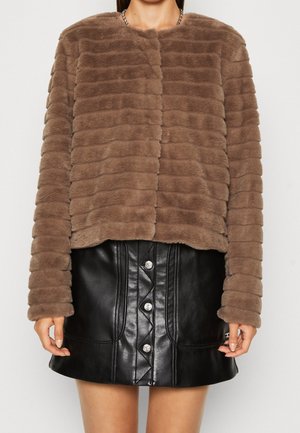 Kobieta ubrana w brązową kurtkę z poziomymi paskami z futra ekologicznego oraz czarną mini spódniczkę z guzikami, wykonaną ze skóry, stojąca na jednolitym tle.