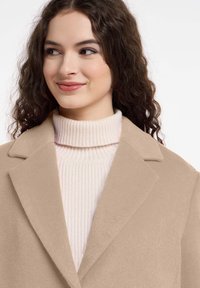 Beige wollen jas met notch-lapels over een lichtroze ribgebreide coltrui. Gladde textuur, op maat gemaakte vorm en zachte kleurenpalet.