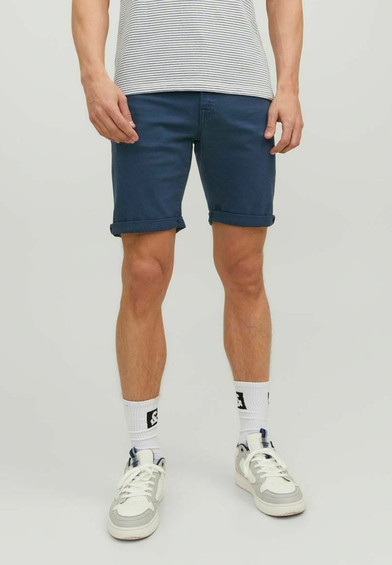Pantaloni corti in cotone blu navy con risvolti, abbinati a una camicia a righe e sneakers bianche. Presentano cuciture a vista e una vestibilità casual.