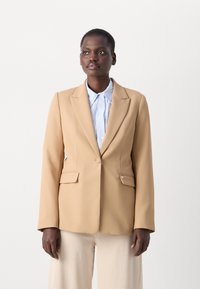 Blazer beige en tissu lisse, avec une fermeture à un bouton, des revers crantés et deux poches avant. Porté par-dessus une chemise bleu clair.