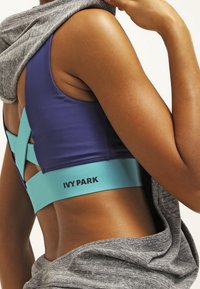 Sujeción deportiva morado con una banda teal que presenta el logo "IVY PARK"; sudadera gris colgada de manera casual sobre el hombro; textura suave y diseño ajustado.